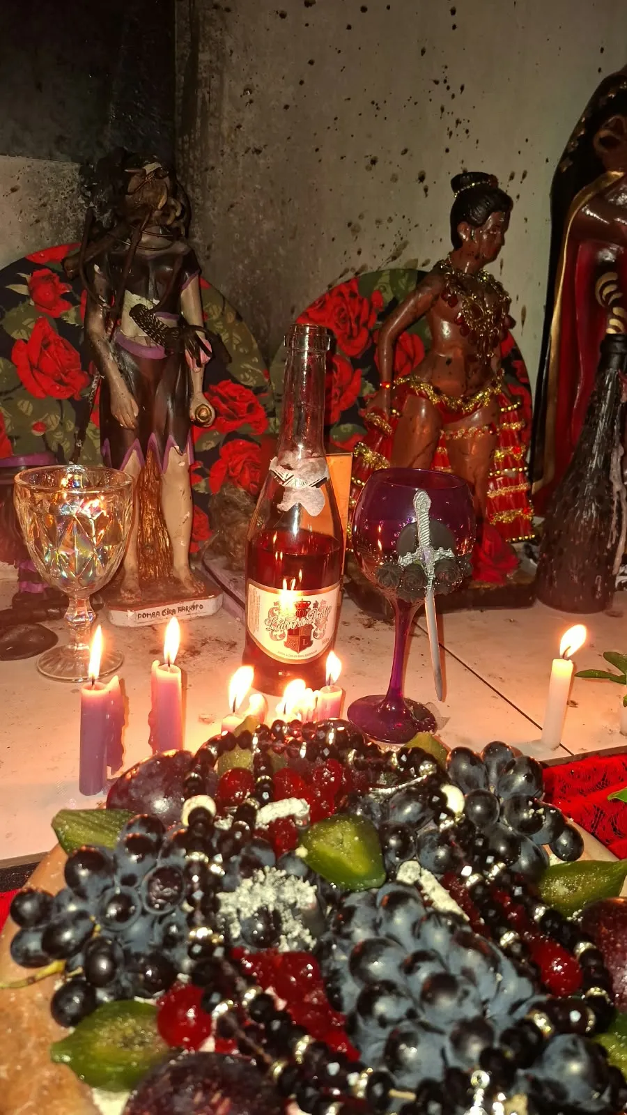 Assentamento à luz de velas roxas e brancas, com imagens de Pombagira, taças, espumante e uvas pretas diante da parede marcada de fuligem