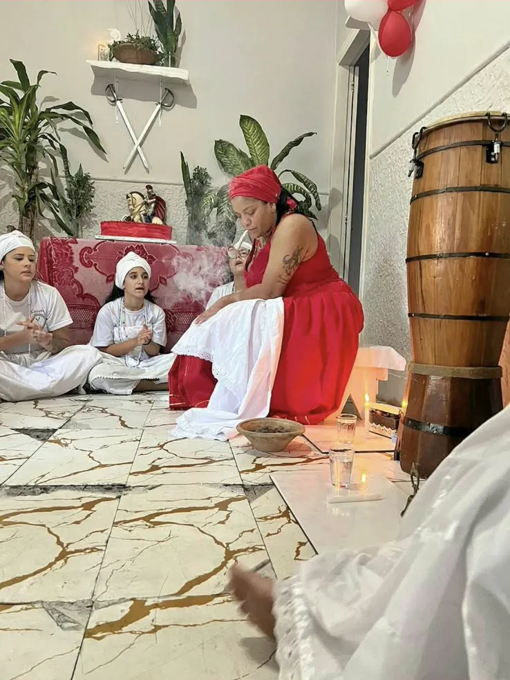 Médium de vestido vermelho e turbante sentada no chão do terreiro, com filhos da casa batendo palmas e um atabaque ao lado